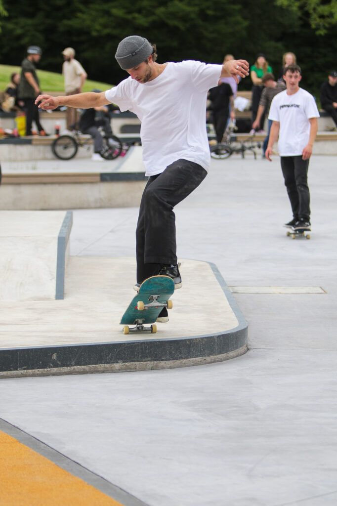 Ein Skateboarder führt einen Trick auf einem Skateboard in einem Skatepark aus. Der Skateboarder befindet sich mitten in einem Manöver, mit einem Fuß auf dem Skateboard und dem anderen ausgestreckt, um das Gleichgewicht zu halten. Im Hintergrund sind andere Personen zu sehen, die anscheinend skaten oder zuschauen. Der Skatepark verfügt über verschiedene Rampen und Schienen, die für Skateboard-Tricks konzipiert sind. Dieses Bild ist interessant, da es das Geschick und das Gleichgewicht beim Skateboarden einfängt und eine aktive Outdoor-Sport- und Freizeitaktivität hervorhebt. 