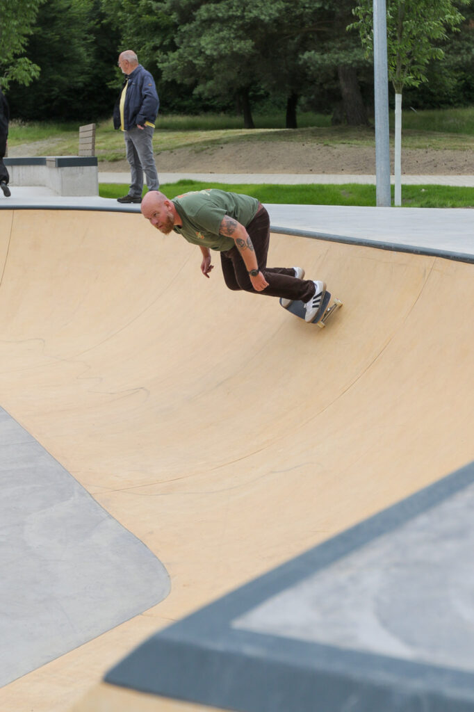 Ein Skateboarder führt auf einer Rampe in einem Skatepark einen Trick aus. Der Skateboarder ist nach unten gebeugt, während er die gekrümmte Oberfläche der Rampe entlangfährt. Im Hintergrund sind Bäume und eine andere Person zu sehen, die zuschaut. Dieses Bild könnte interessant oder relevant sein für Diskussionen über Skateboarding, Outdoor-Aktivitäten oder Sport im Allgemeinen.
