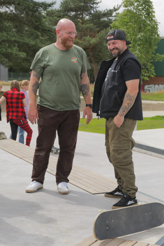 Zwei Personen stehen auf einer Holzrampe in einem Skatepark. Eine trägt ein grünes T-Shirt und braune Hosen, die andere ein schwarzes T-Shirt mit dunklen Hosen. Ein Skateboard liegt seitlich vor ihnen auf der Rampe. Die Szene scheint im Freien zu sein, mit Bäumen und bewölktem Himmel im Hintergrund sowie einer weiteren Person, die in der Ferne teilweise sichtbar ist. Dieses Bild könnte relevant sein für Diskussionen über Skateboarding, Outdoor-Aktivitäten oder soziale Interaktionen an öffentlichen Orten wie Skateparks.