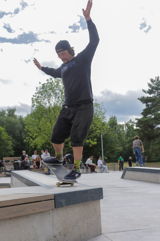 Eine Person wird mitten in der Aktion beim Skateboardfahren auf einer Kante in einem Skatepark erfasst. Die Person trägt ein langärmeliges schwarzes Hemd, schwarze Shorts und eine Kappe. Das Skateboard ist auf seinen Vorderrädern geneigt, während die Person die Arme zur Stabilität ausstreckt. Im Hintergrund sind andere Personen zu sehen, die als Zuschauer erscheinen oder auf ihre Reihe warten, Bäume und ein bewölkter Himmel, der auf eine Außenumgebung bei Tageslicht hindeutet.