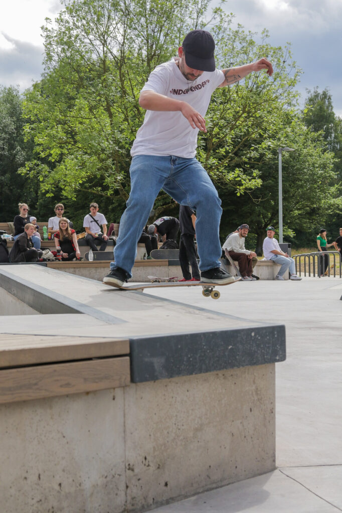 Das Bild zeigt eine Person beim Ausführen eines Skateboard-Tricks in einem Skatepark im Freien. Dieser Moment ist interessant, da er das Können und Timing zeigt, das für die Ausführung solcher Manöver erforderlich ist. Die Anwesenheit eines Publikums trägt zur dynamischen Atmosphäre der Umgebung bei.