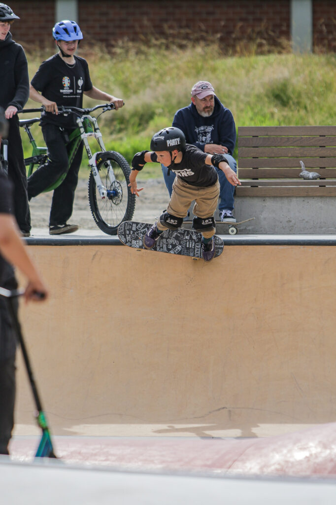 Eine Person führt auf einem Skateboard am Rand einer Skate-Bowl einen Trick aus. Der Skateboarder ist nach unten gebeugt, während er den Trick ausführt. Im Hintergrund sind zwei Personen zu sehen, eine davon steht mit einem Fahrrad und die andere hält teilweise sichtbar, was wie ein Roller aussieht. Die Szene spielt sich in einem Skatepark im Freien ab, mit Betonstrukturen, die für Skate-Aktivitäten konzipiert sind. Dieses Bild ist interessant, da es die dynamische Bewegung und das Können beim Skateboarden einfängt und einen actionreichen Moment im Skatepark hervorhebt.