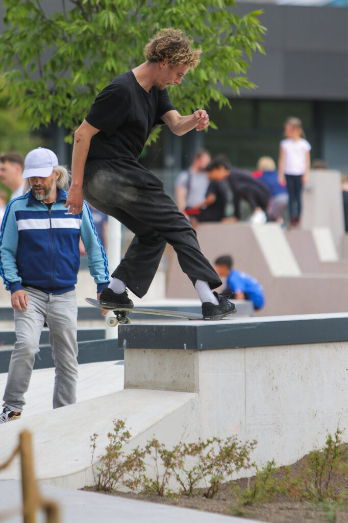 Eine Person führt einen Trick auf einem Skateboard aus, indem sie auf der Kante einer Betonkante in einem Skatepark im Freien entlangrutscht. Im Hintergrund sind mehrere andere Personen, die wahrscheinlich zuschauen oder auf ihre Reihe warten. Der Fokus liegt auf der Aktion des Skateboarders, und die urbane Umgebung des Skateparks trägt zur dynamischen Atmosphäre des Bildes bei.