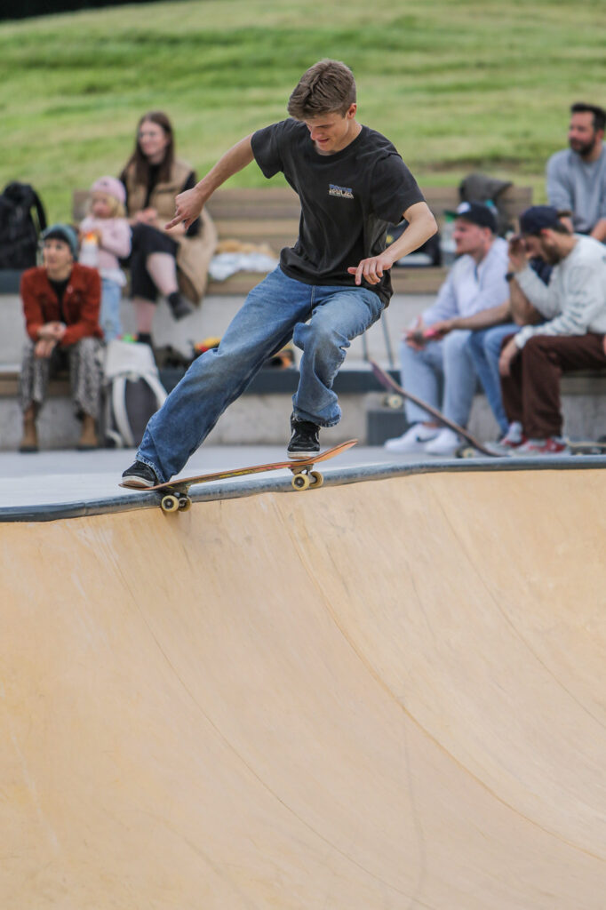 Ein Skateboarder in einem grauen T-Shirt und blauen Jeans fährt auf der Kante einer Halfpipe in einem Skatepark. Er balanciert das Skateboard und konzentriert sich auf seinen Trick. Im Hintergrund sitzen und stehen mehrere Personen auf einer Bank und beobachten ihn. Die Umgebung ist grün und scheint in einem Park zu sein.
