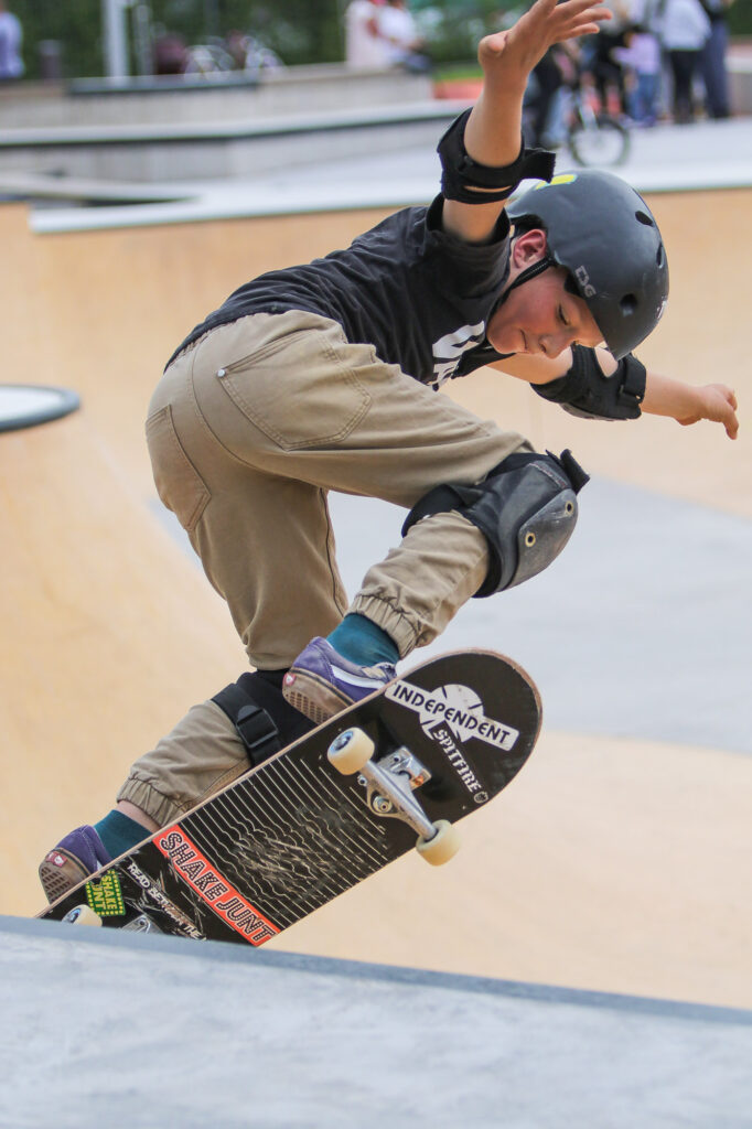 Ein Junge mit Schutzhelm und Schutzausrüstung fährt Skateboard in einem Skatepark. Er führt einen Trick aus, indem er das vordere Ende des Boards nach oben zieht. Sein linker Arm ist ausgestreckt und sein Gesicht konzentriert. Im Hintergrund sind unscharf andere Personen zu sehen.