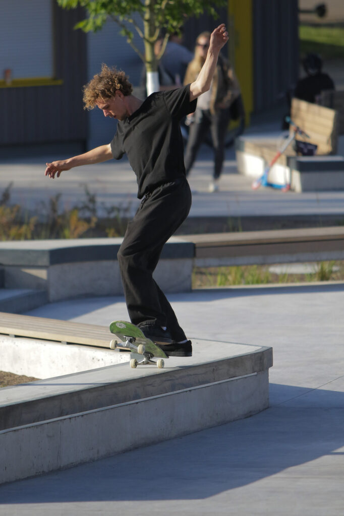 Ein Skater führt einen Trick auf einer erhöhten Betonfläche im Skatepark aus. Im Hintergrund sind Zuschauer und ein paar Bäume zu sehen, ebenso wie eine moderne Parklandschaft mit Sitzbänken. Das Wetter ist sonnig, und einige Menschen schauen dem Skater zu.