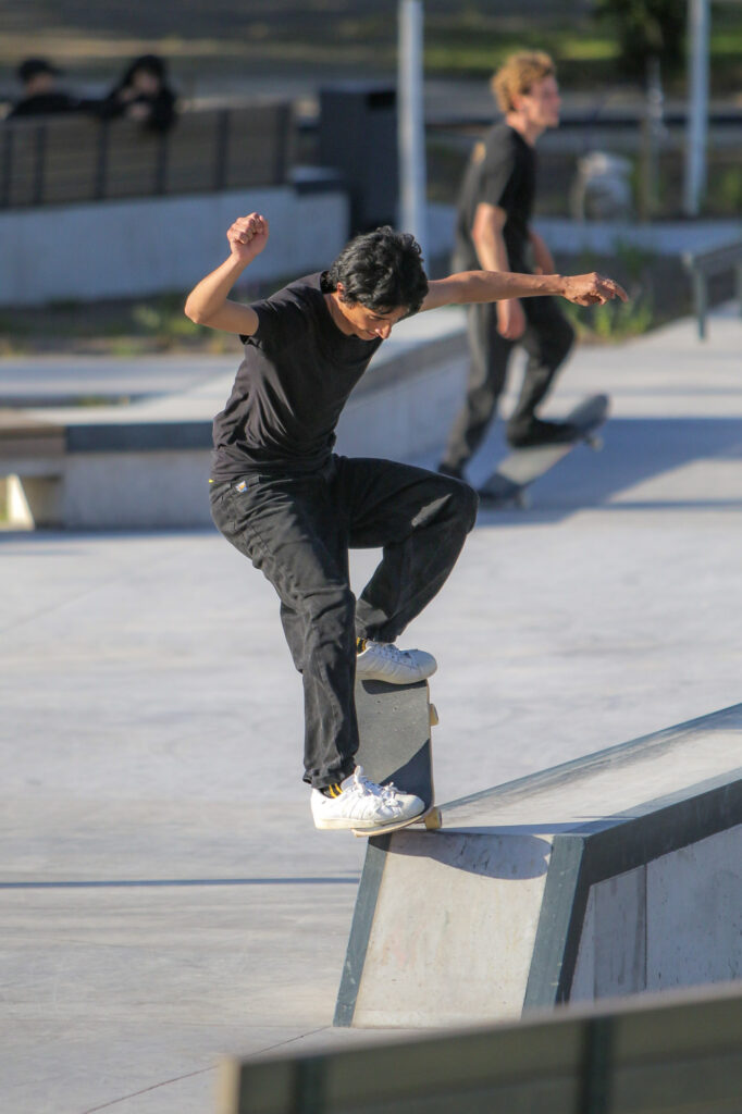 Ein Skater führt einen Trick auf einer Kante im Skatepark aus. Im Hintergrund ist ein weiterer Skater zu sehen, der ebenfalls auf dem Skateboard steht. Die Szene spielt sich bei sonnigem Wetter ab, und im Hintergrund sind einige Zuschauer an einem Geländer zu erkennen.