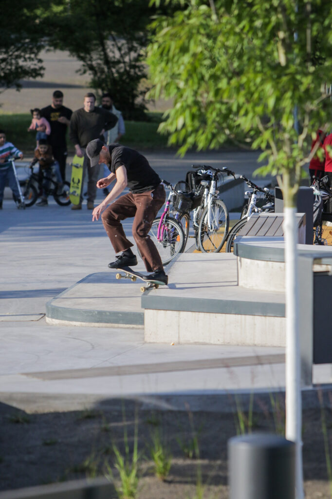 Ein Skater führt einen Trick auf einer Rampe in einem Skatepark aus. Im Hintergrund stehen mehrere Menschen, darunter Kinder mit Fahrrädern und einem Skateboard. Die Szene ist von Bäumen und Grünflächen umgeben, und das Wetter scheint sonnig zu sein.